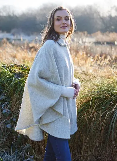 Side shot of woman standing against wintery countryside backdrop wearing cream wool cape. 